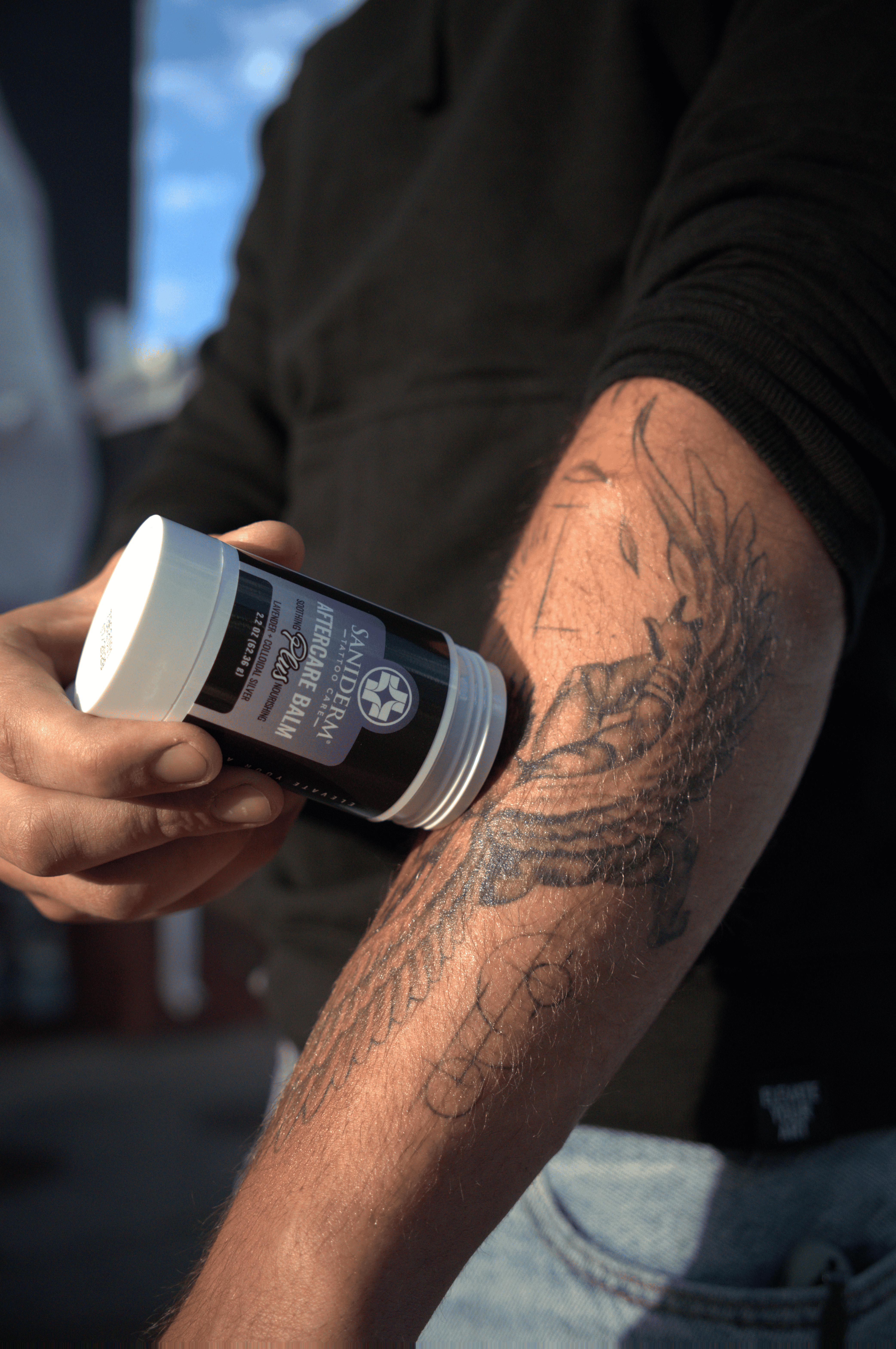 A person holding a white and black container of tattoo balm against their tattooed forearm, with the product being applied or demonstrated in a shop setting.